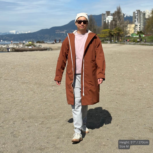 A male swimmer wears a Tingting swim parka sPARKA Fly in brown at the beach in Vancouver. He styles it with a white beanie, sun glasses, light pink t shirt, gray sweat pants, and ivory sneakers.